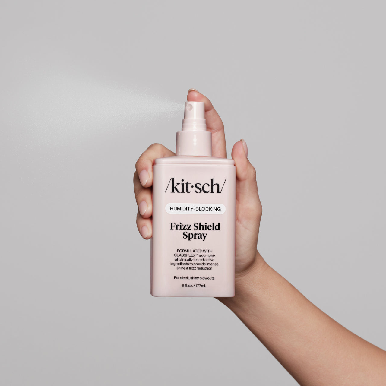 KITSCH Humidity Blocking Frizz Shield Spray — hand holding bottle from below with nozzle up, spray mist visible — product action shot on light gray background — Humidity-Blocking | Frizz Shield Spray — Formulated with GLASSPLEX™ — For sleek, shiny blowouts — 6 fl. oz. / 177mL — Hydrates without weighing hair down | Protects hair from heat up to 266°F/130°C