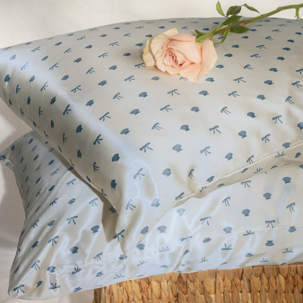 KITSCH Satin Pillowcase in Pearlized Cream & Coastal Blue Print — lifestyle flat lay on wicker surface with pink rose — coastal blue seashells & bows print close-up — Reduces friction against hair & skin, helps reduce breakouts — For hair & skin-loving comfort