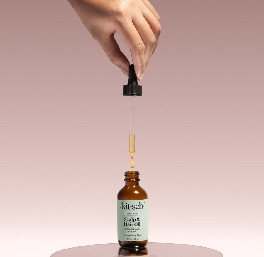 KITSCH Strengthening Rosemary & Biotin Scalp & Hair Oil — product shot — dropper releasing oil into the amber glass bottle on a blush pedestal — 100% plant-based blend of rosemary, jojoba, argan and castor oils with biotin